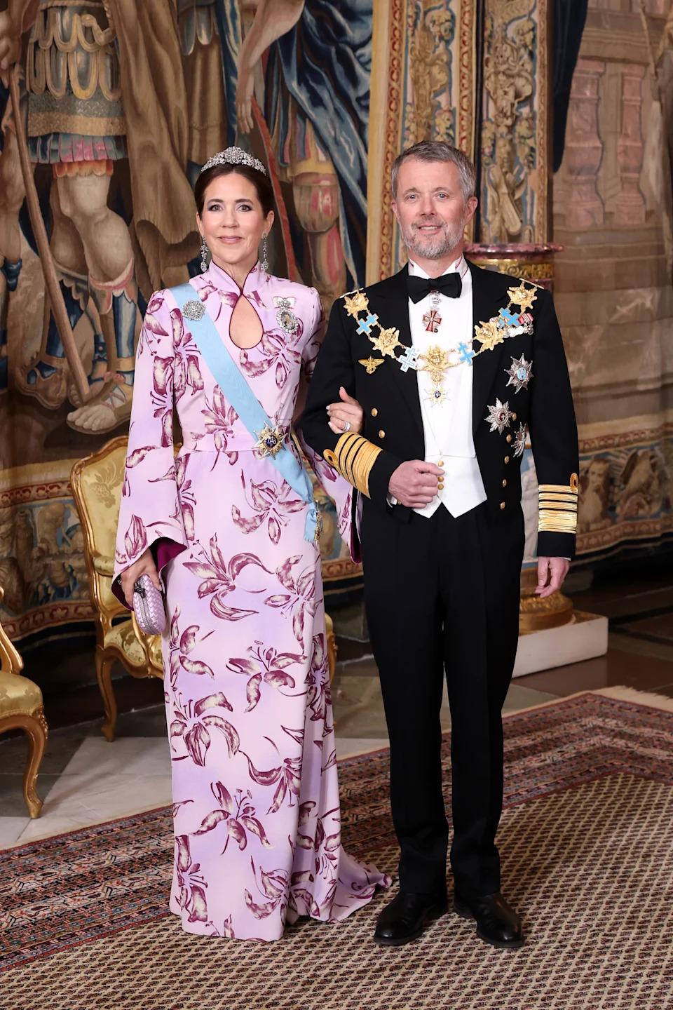 Queen Mary and King Frederik in eveningwear standing in front of a tapestry