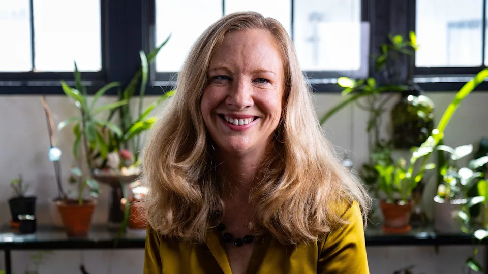 Karen Canfell, wearing a gold top, smiles at the camera, potted plants in the background behind her