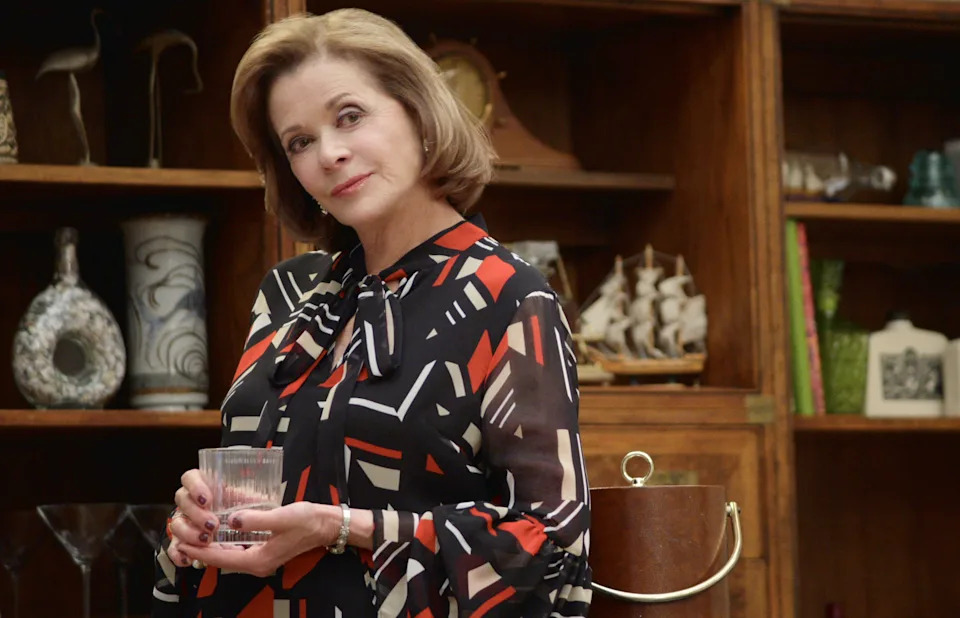 Person holding a glass, wearing a patterned blouse, standing in front of shelves with decor items