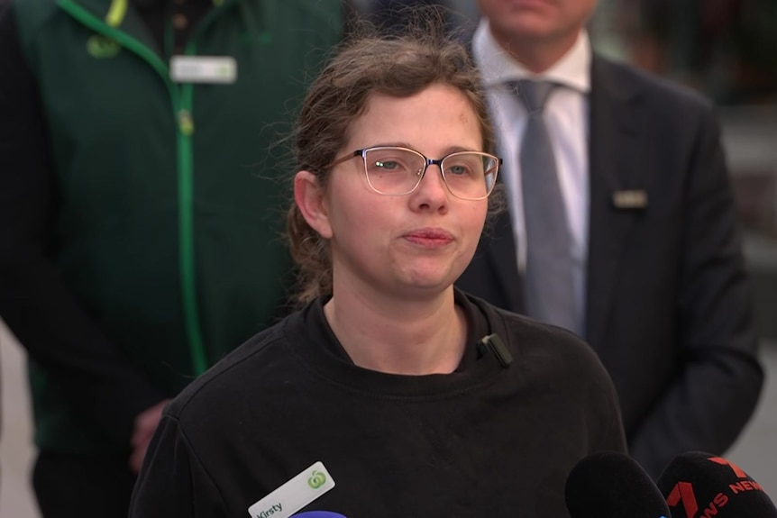 A woman with glasses in a Woolworths uniform stands in front of media microphones and union workers