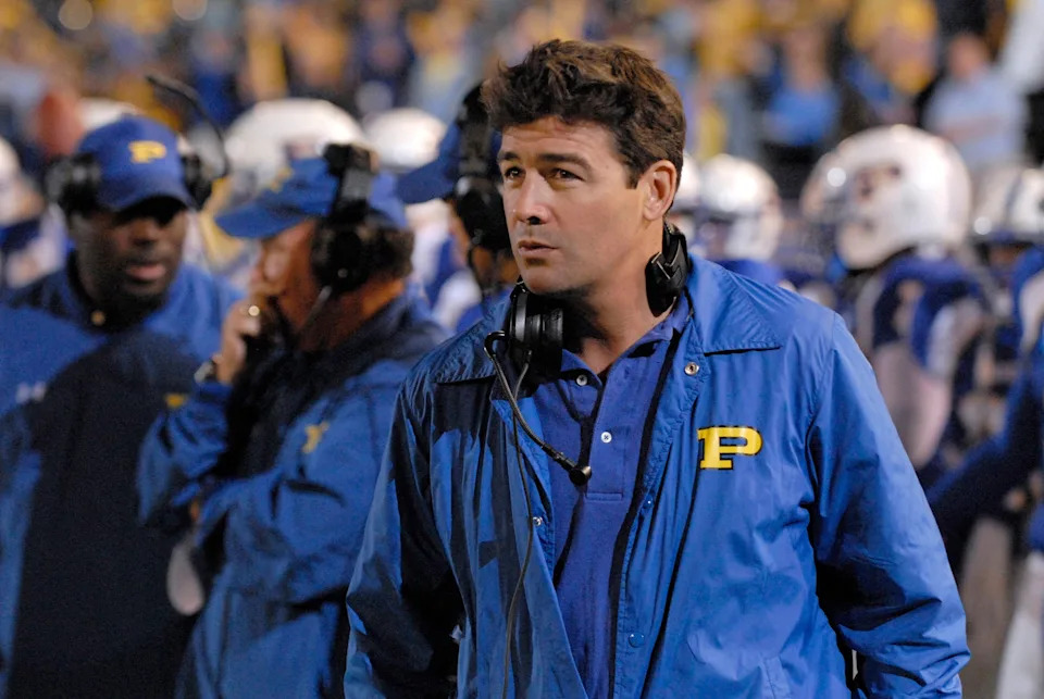 A coach in a blue jacket with headphones stands on a football field, focused, surrounded by a team during a game