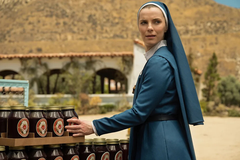 A person dressed as a nun inspects jars on a table outdoors, in a scene from a TV show or movie set