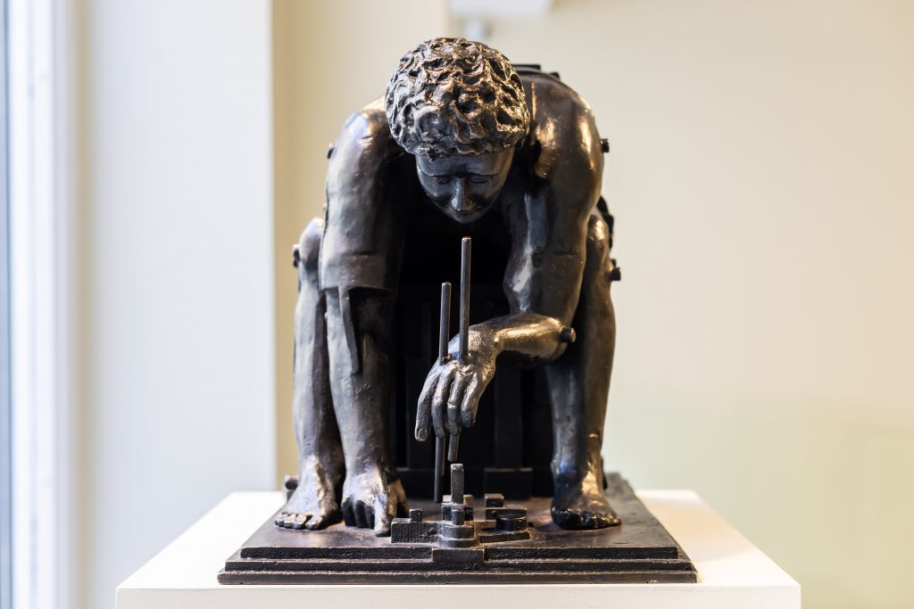 A bronze sculpture of a crouching male figure leaning forward over a small arrangement of geometric forms. The figure’s posture is compact, with bent knees and one hand extended downward, interacting with upright rods and block-like elements on the base. The surface is textured and industrial in feel, and the piece is displayed on a pedestal in a gallery setting. Featured in a British sculpture exhibition at Bowman Sculpture, London.