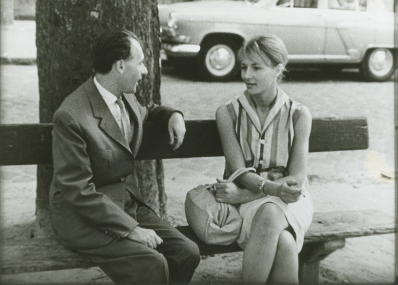 a black-and-white still of a man and woman sitting on a park bench by the street