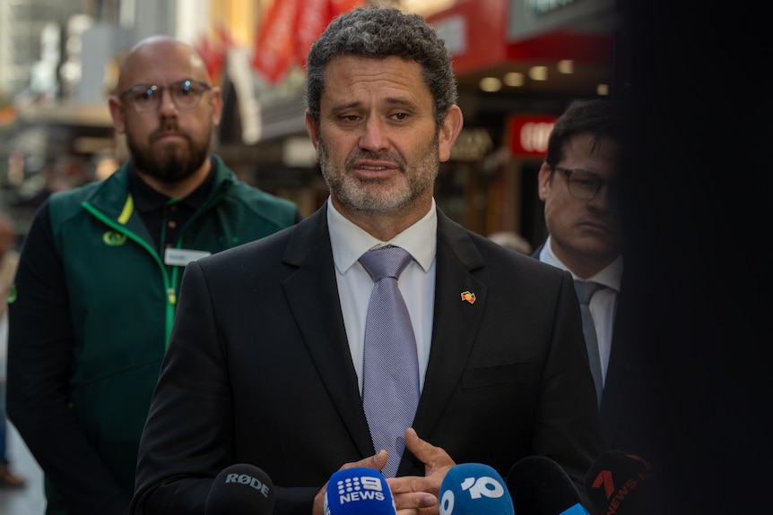 A man in a suit is standing behind media microphones in a shopping mall and speaking.