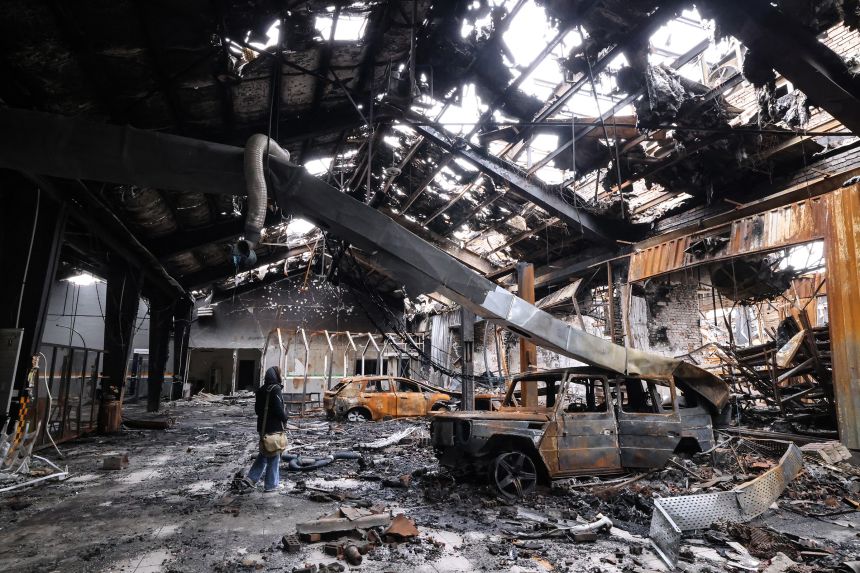 A journalist stands next to the wreckage of a vehicle during the visit to a car service center in eastern Tehran that was hit by a missile strike, on March 28, 2026.