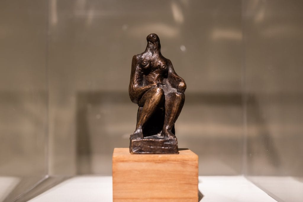 A small bronze sculpture of a seated human figure displayed on a wooden plinth inside a glass case. The figure is stylized and somewhat abstract, with elongated limbs and a simplified head, sitting with one knee raised and arms resting on its legs. The background is softly blurred, emphasizing the sculpture. Featured in a British sculpture exhibition at Bowman Sculpture, London.