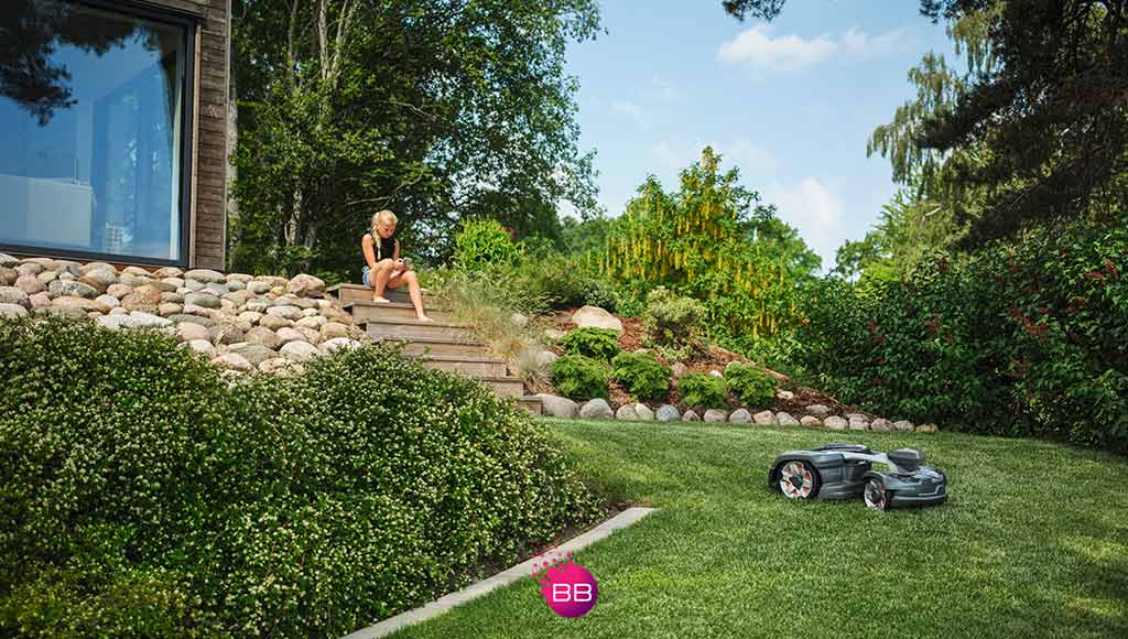 A child sitting on the steps of a house with a large green space in front of her and a Husqvarna lawnmower working