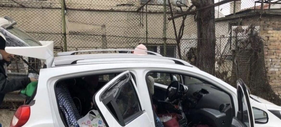 A white hatchback car is packed with belongings and luggage, with its doors open. A person is seen at the rear of the vehicle, appearing to be in the process of loading or unloading items.