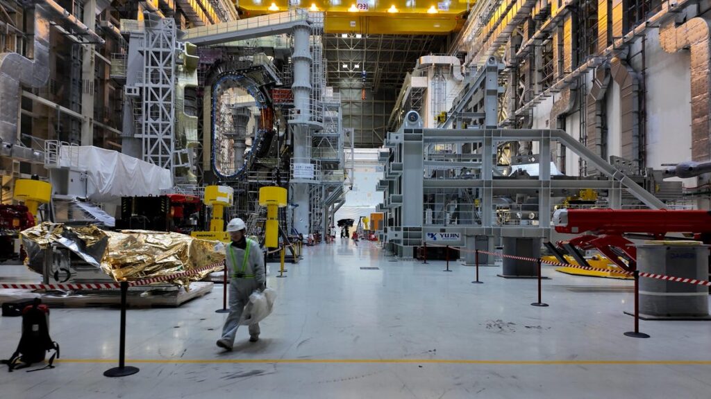 Inside the World’s Biggest Bet on Fusion Energy inside iter's tokamak assembly hall a high-cieling building filled with tokamak components and a worker walking by