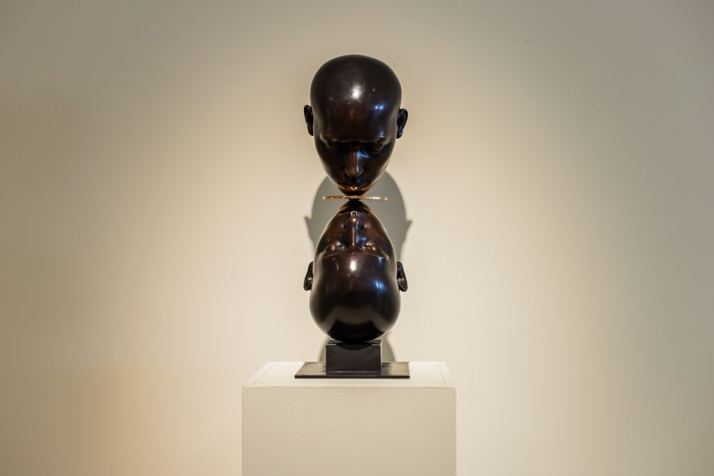 A polished dark bronze sculpture of two human heads stacked vertically on a pedestal, one upright and one inverted, their lips nearly touching at a thin horizontal disc between them. The minimalist gallery setting and focused lighting emphasize the smooth surface and reflective quality of the forms. Featured in a British sculpture exhibition at Bowman Sculpture, London.