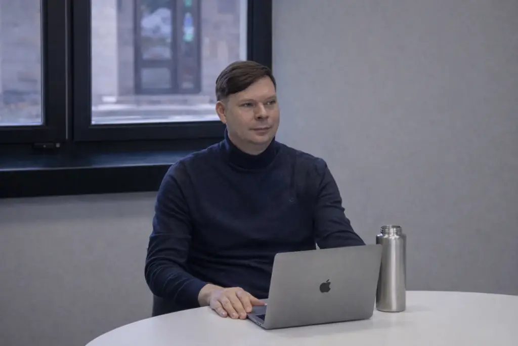 AI SEO in Lithuania: What Businesses Should Know Vyras tamsiai mėlynu megztiniu aukštu kaklu sėdi prie stalo su nešiojamuoju kompiuteriu ir gertuve.