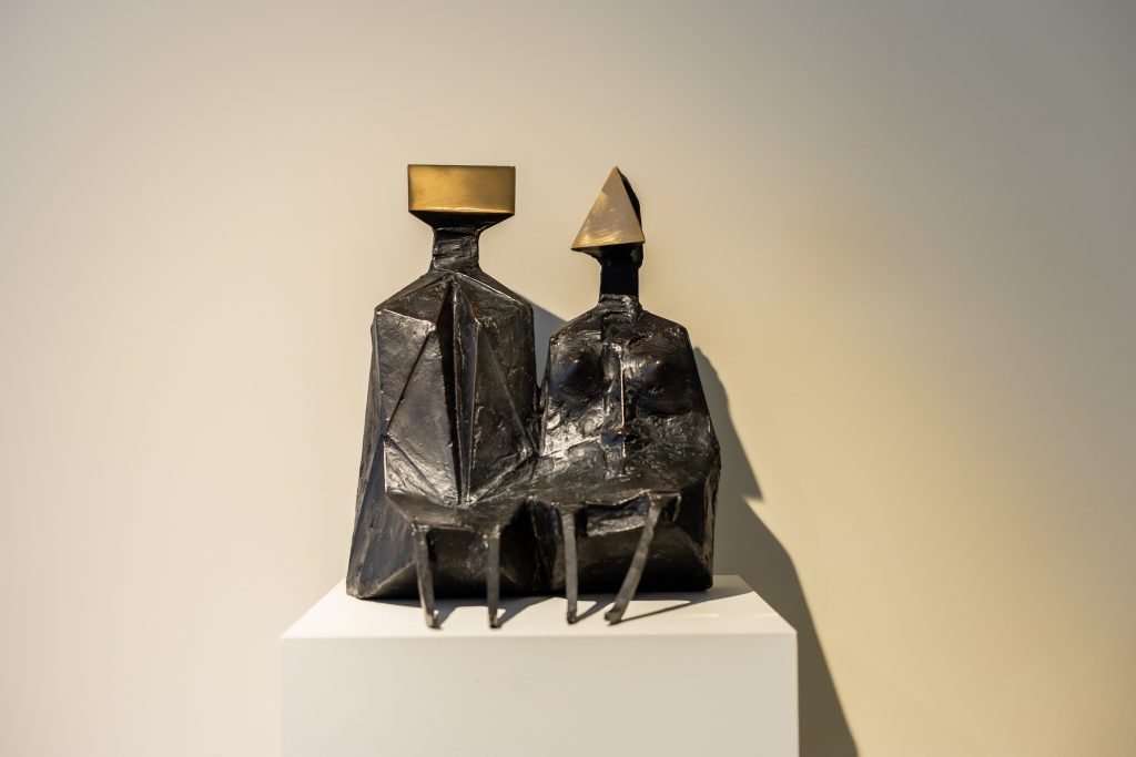 An abstract bronze sculpture of two seated human figures on a pedestal, each with angular, faceted bodies and geometric heads—one topped with a rectangular form and the other with a triangular shape. Their elongated legs extend forward, and the dark, textured surface contrasts with the smooth, light-colored gallery wall behind them.