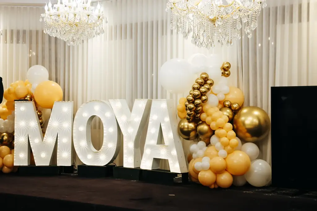 They forgot the “North”: Greek priests and Australian politicians at Skopjan event Illuminated letters spell 'MOYA' on stage, surrounded by gold, white, and cream balloons at an elegant event backdrop.
