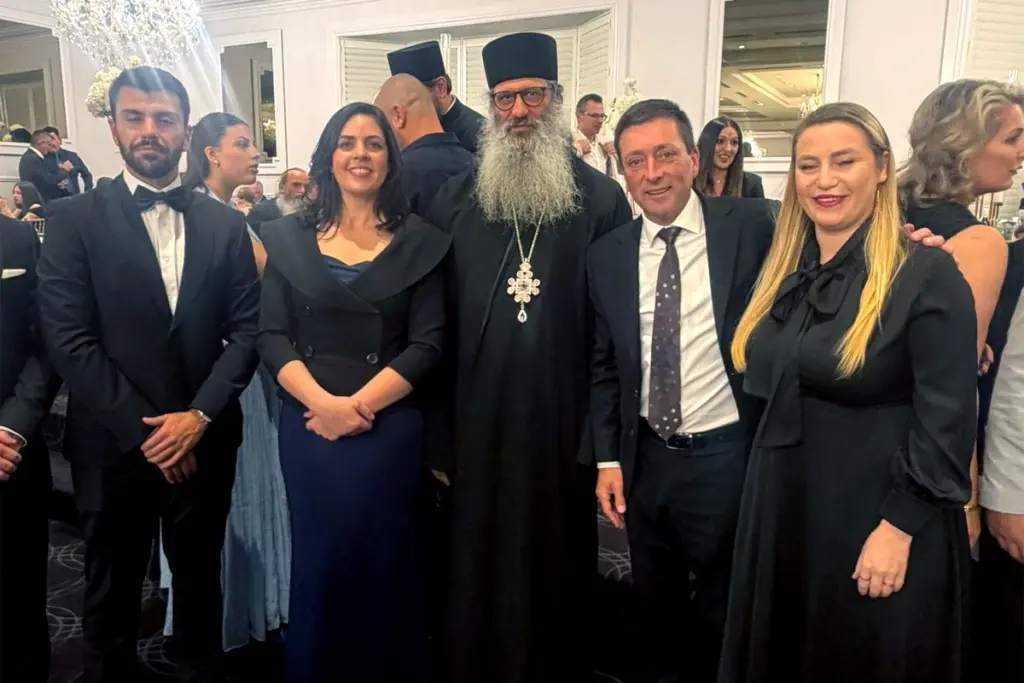 Did they forget the “North”? Group of five adults in formal attire posing at a gala, with an Orthodox priest wearing a black robe and cross in the center