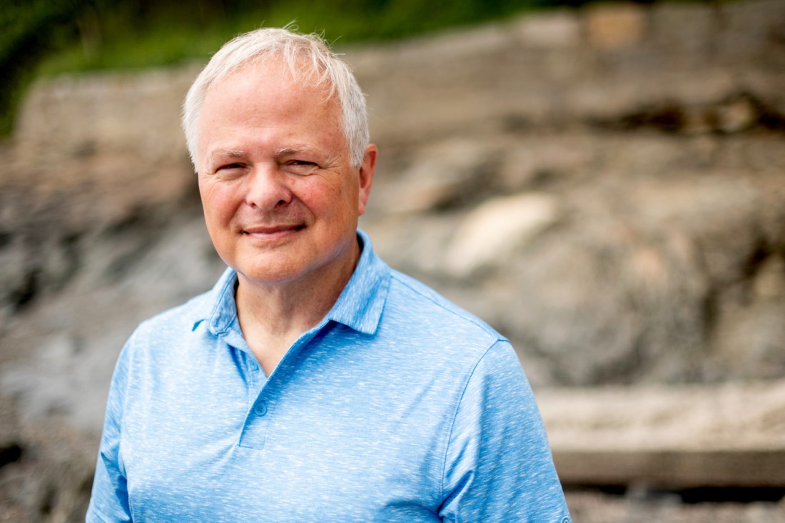 Mark Patterson is an older white man. He is wearing a light blue collared shirt and is standing against a rocky backdrop.