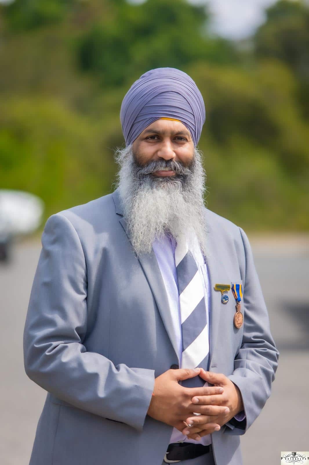 A Sikh man wearing a turban and a suit stands with his hands clasped in front.