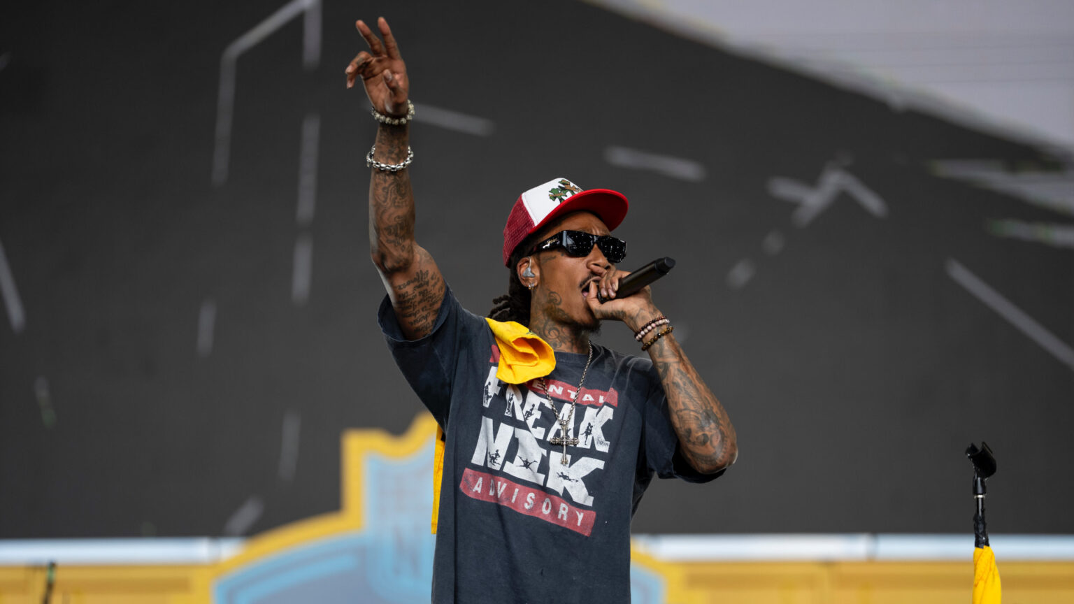 Wiz Khalifa wearing a cap, sunglasses, and a graphic t-shirt holds a microphone on stage, gesturing with one hand.