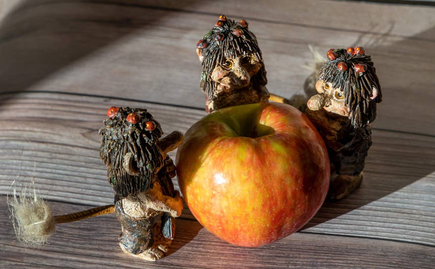 Handcrafted Icelandic troll figurines gathered playfully around a large red apple on a wooden surface. Handcrafted Icelandic troll figurines gathered playfully around a large red apple on a wooden surface.