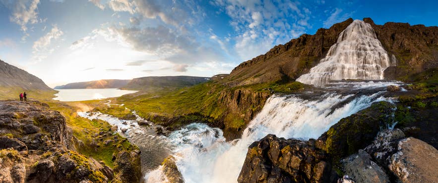 Panoramic view of Dynjandi Waterfall in Iceland cascading down cliffs into a river, with hikers and fjord scenery at sunset. Panoramic view of Dynjandi Waterfall in Iceland cascading down cliffs into a river, with hikers and fjord scenery at sunset.