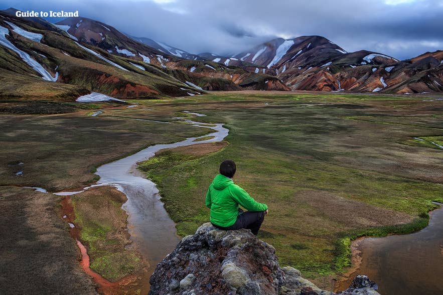 A person in a green jacket sits on a rock overlooking the colorful mountains of Landmannalaugar, Iceland. A person in a green jacket sits on a rock overlooking the colorful mountains of Landmannalaugar, Iceland.
