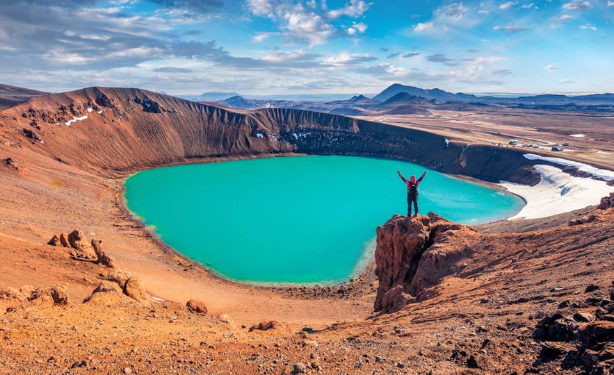 Hiker stands by the vivid turquoise Viti Crater Lake in Iceland’s Krafla volcanic area under a partly cloudy sky. Hiker stands by the vivid turquoise Viti Crater Lake in Iceland’s Krafla volcanic area under a partly cloudy sky.