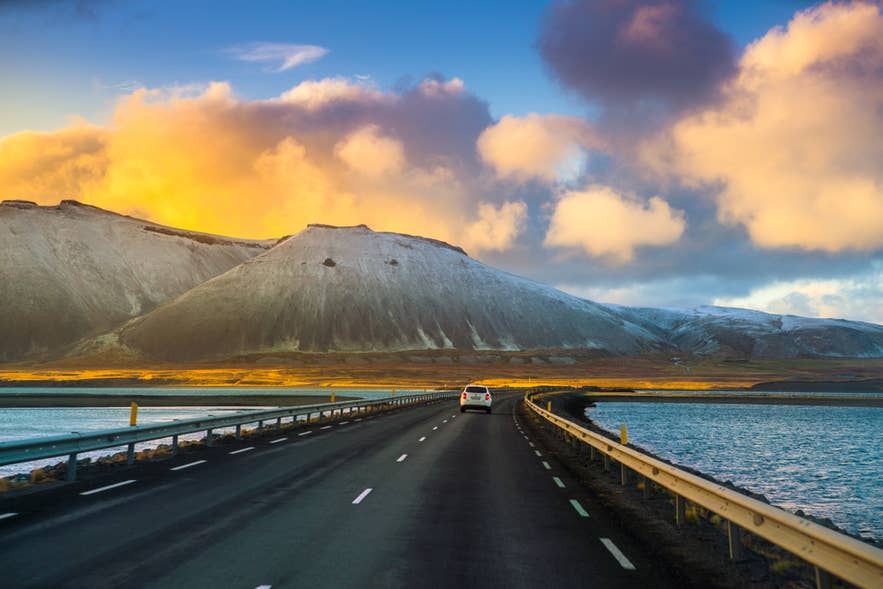 Car drives on Iceland's Ring Road beside water and snowy mountains at sunset, with vibrant clouds lighting the sky. Car drives on Iceland's Ring Road beside water and snowy mountains at sunset, with vibrant clouds lighting the sky.