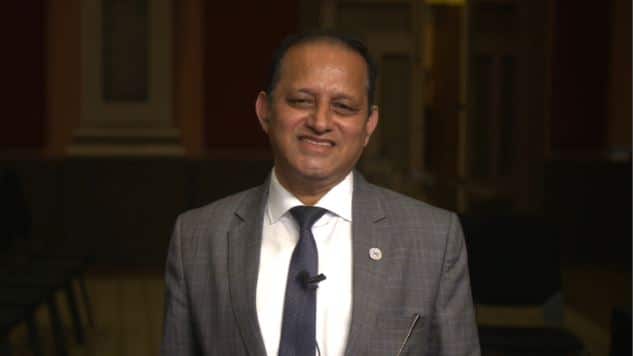 A man in a grey suit and matching tie smiles.