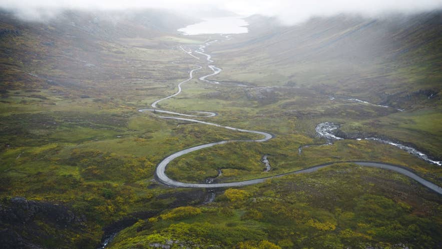Winding mountain road cuts through a green Icelandic valley, leading toward a misty lake under low clouds.