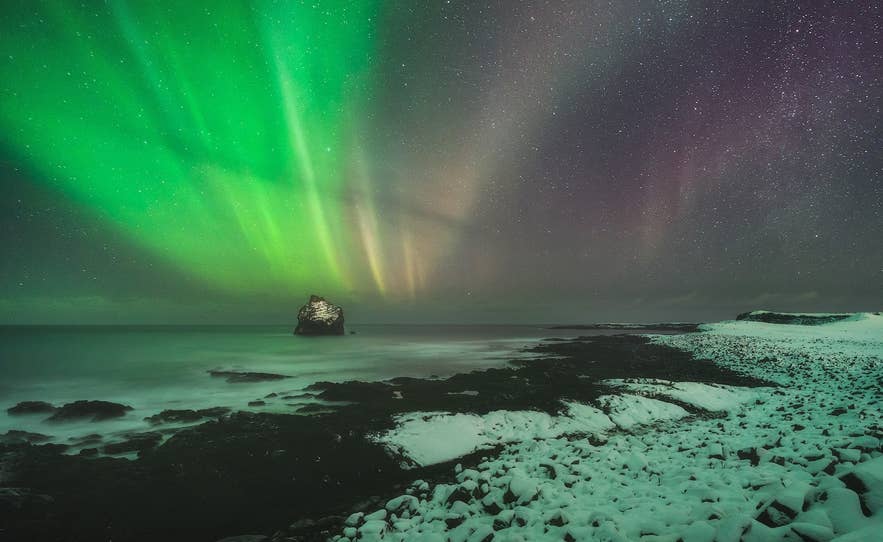 Northern Lights glow above a snowy Icelandic coastline, with vivid green auroras lighting the night sky over the ocean.
