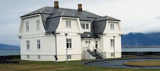 The white wooden Hofdi House stands by the sea with a gray roof and mountains in the background. The white wooden Hofdi House stands by the sea with a gray roof and mountains in the background.