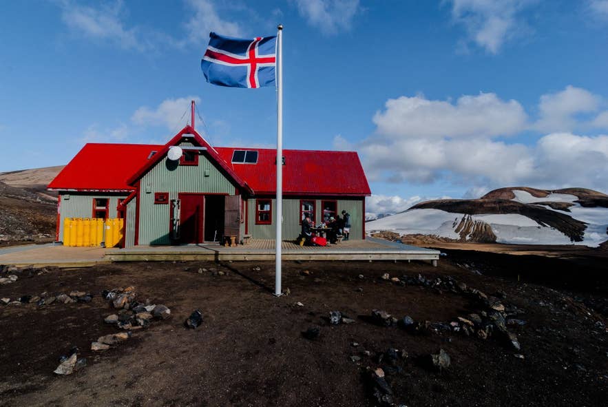 Icelandic flag waves in front of a red-roofed mountain hut with snowy hills and rocky ground under a bright blue sky. Icelandic flag waves in front of a red-roofed mountain hut with snowy hills and rocky ground under a bright blue sky.