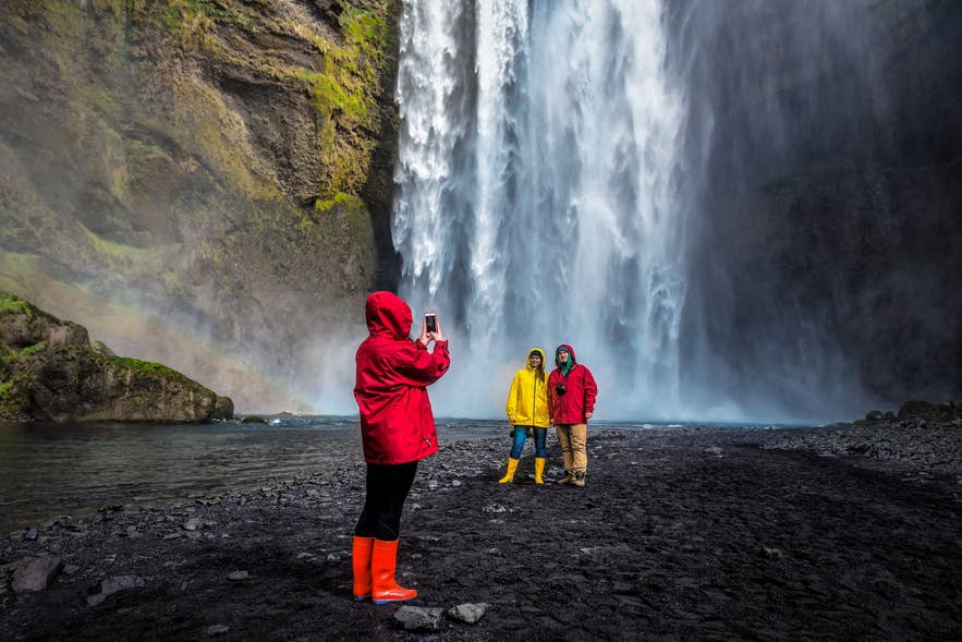 Visitors in colorful raincoats taking photos at Skogafoss Waterfall in Iceland with mist and rainbow