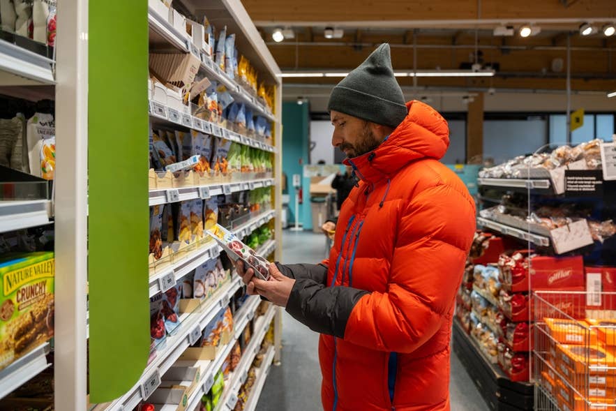 A shopper in an orange jacket browsing grocery shelves at a Reykjavik supermarket.