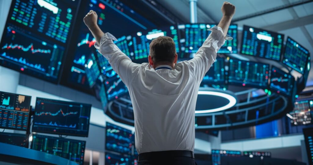 AI Supercharged This Flatlining U.S. Manufacturing Stock. Now It’s Just Scored a Massive New Nvidia Partnership. A person facing away from the viewer raises their fists in triumph as they watch a set of stock price monitors.