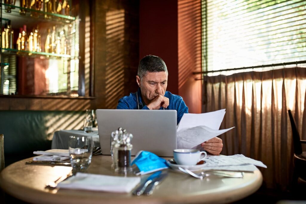 An investor sits at a table in a cafe while looking at some papers and a laptop computer.