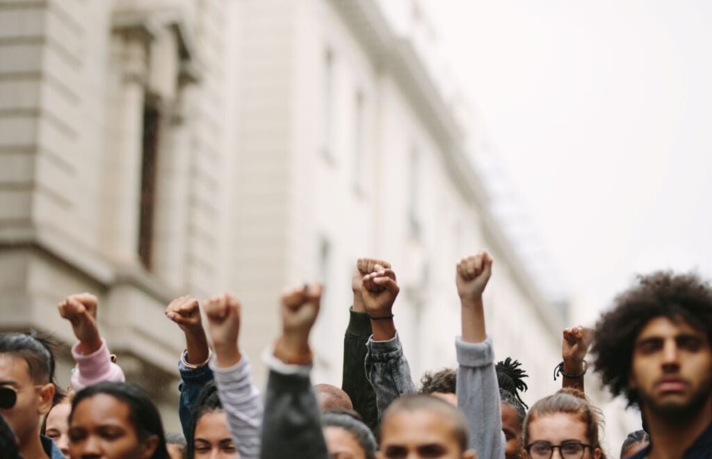 A group of people in front of a large white building hold up their fists in protest.