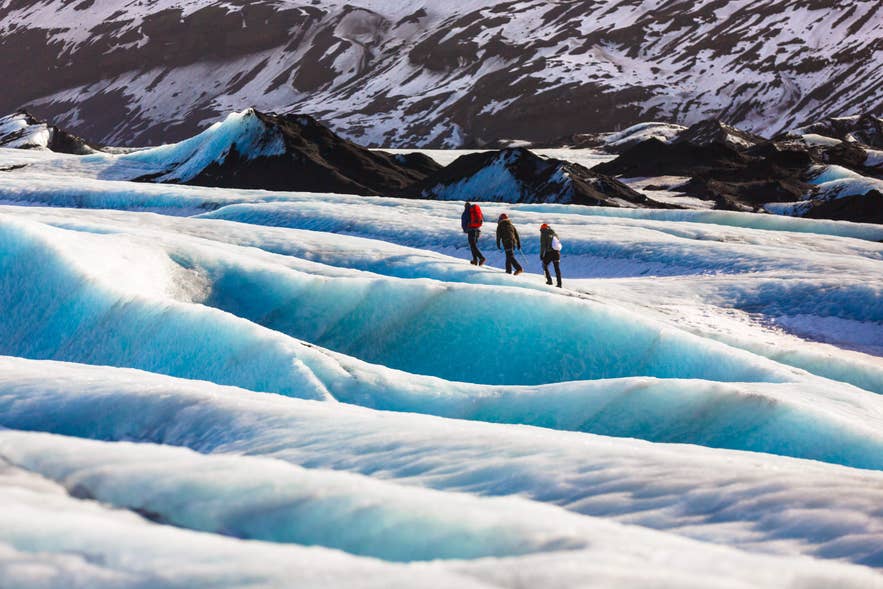 Hikers walk across bright blue ice on a glacier in Iceland, surrounded by ridges and snow-covered mountains.