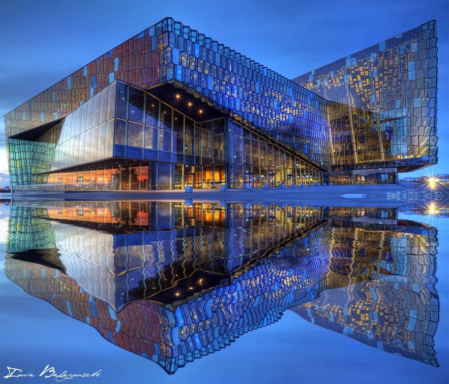 Harpa Concert Hall in Reykjavik glowing under a bright spring twilight sky, with its glass facade reflected in still water.