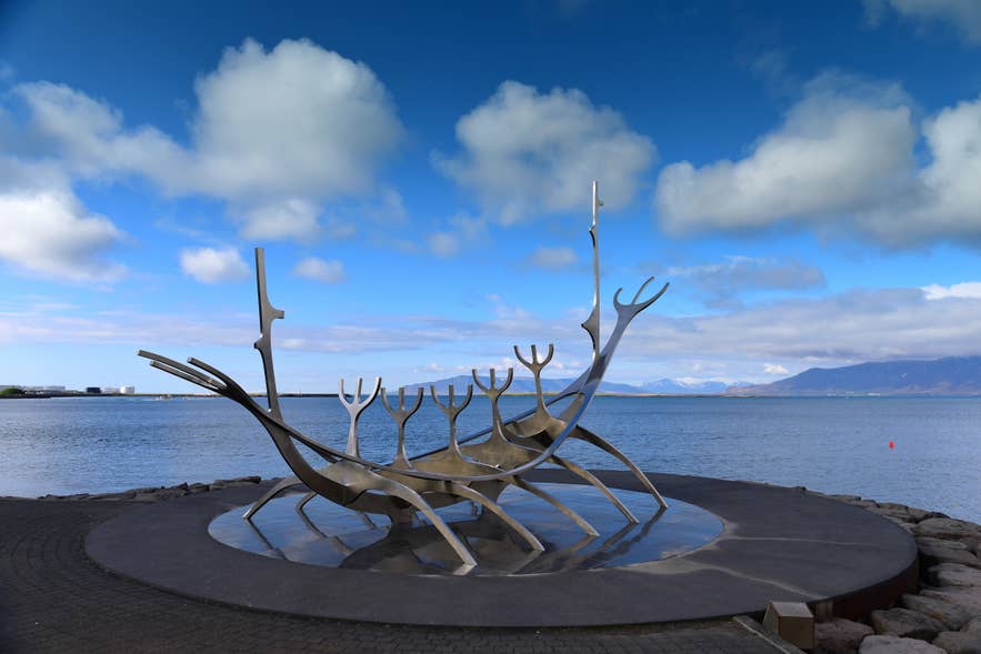 The Sun Voyager sculpture stands by the Reykjavik waterfront under a bright sky with mountains visible across the bay.