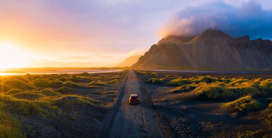 A car driving along a remote gravel road in Iceland at sunset, surrounded by mossy lava fields and dramatic mountains.