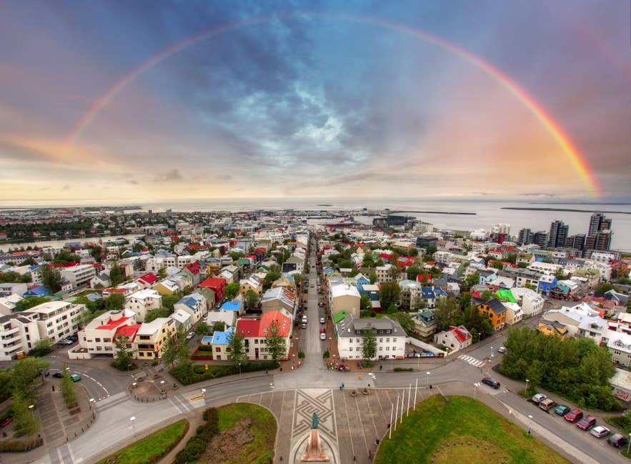 A rainbow arcs over central Reykjavik, showcasing colorful houses, city streets, and the coastline under a bright spring sky.
