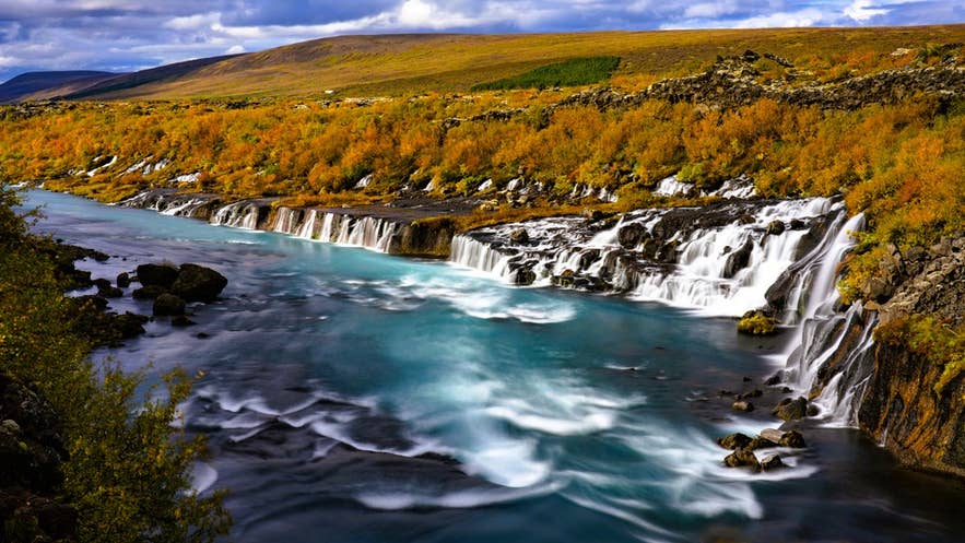 Hraunfossar Waterfall in autumn surrounded by golden foliage, a scenic stop where layers are key for what to wear in Iceland. Hraunfossar Waterfall in autumn surrounded by golden foliage, a scenic stop where layers are key for what to wear in Iceland.