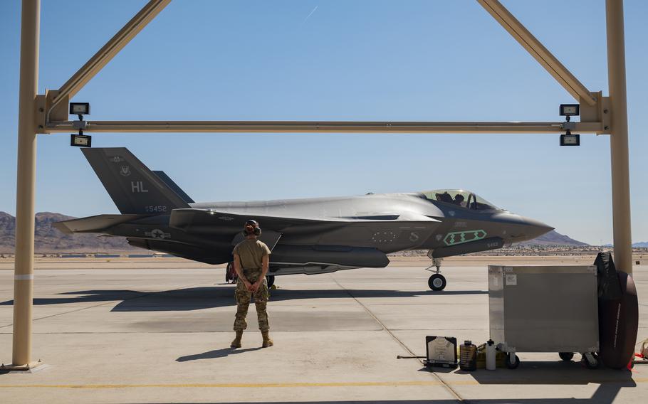 An F-35A Lightning II fighter jet sits on a sunlit flight line with a crew chief in uniform standing at parade rest in front of the aircraft.
