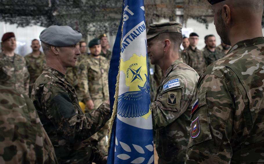 Two soldiers in camouflage uniforms shake hands across a blue NATO battalion flag during a military ceremony, with rows of multinational troops standing in the background.