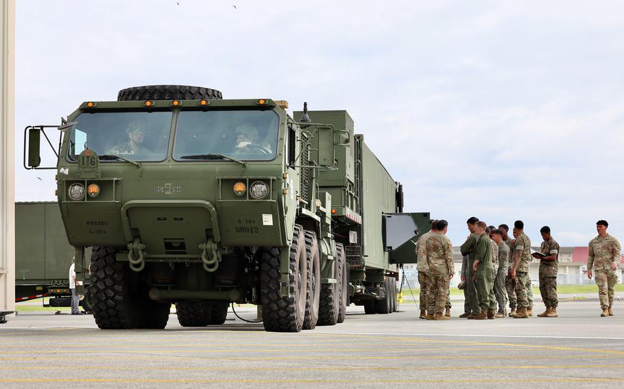 Large green military cargo truck with multiple axles parked on an airfield as a group of uniformed personnel stands nearby.