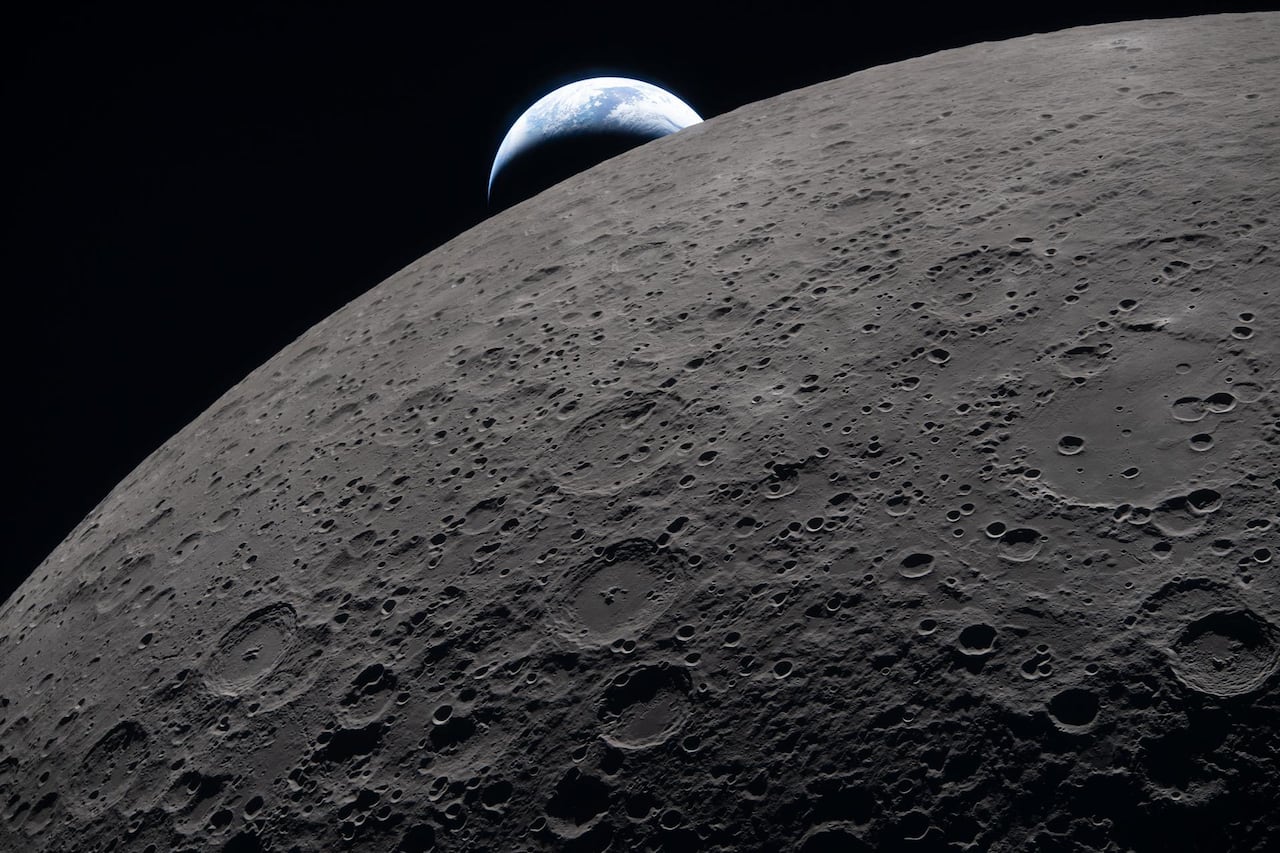 A heavily cratered moon is seen in the foreground with a small crescent Earth in the background.