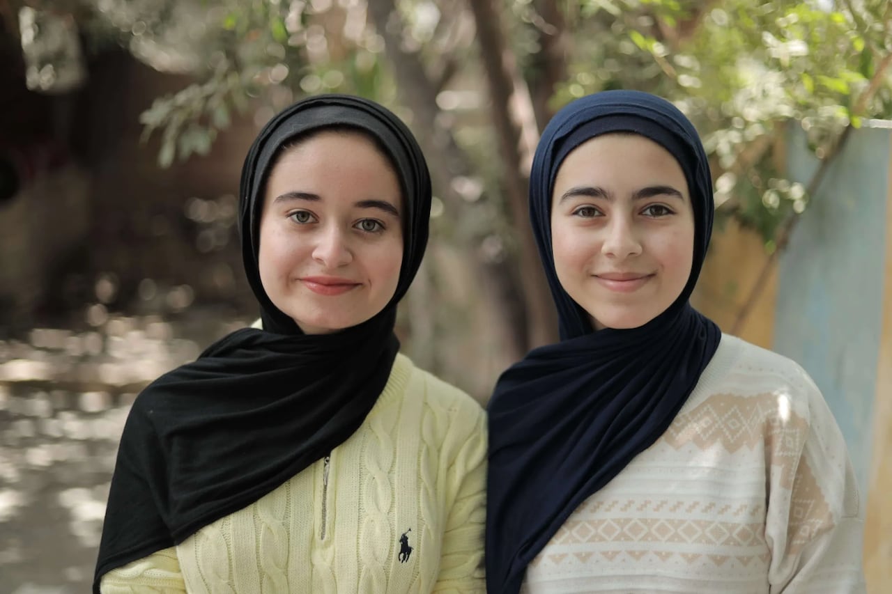 Two girls wearing black headscarves and light sweaters facing the camera with slight smiles