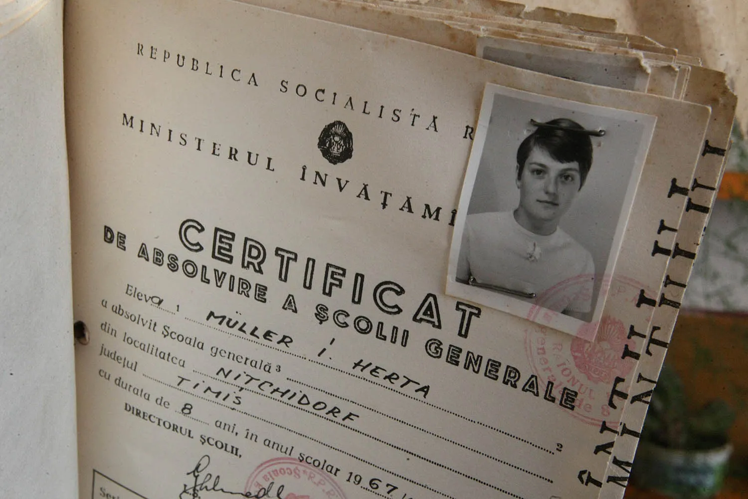 A close-up shot of an open paper document featuring stamped and handwritten text. A black-and-white portrait photo of a person with short hair is stapled to the upper right corner of the page.