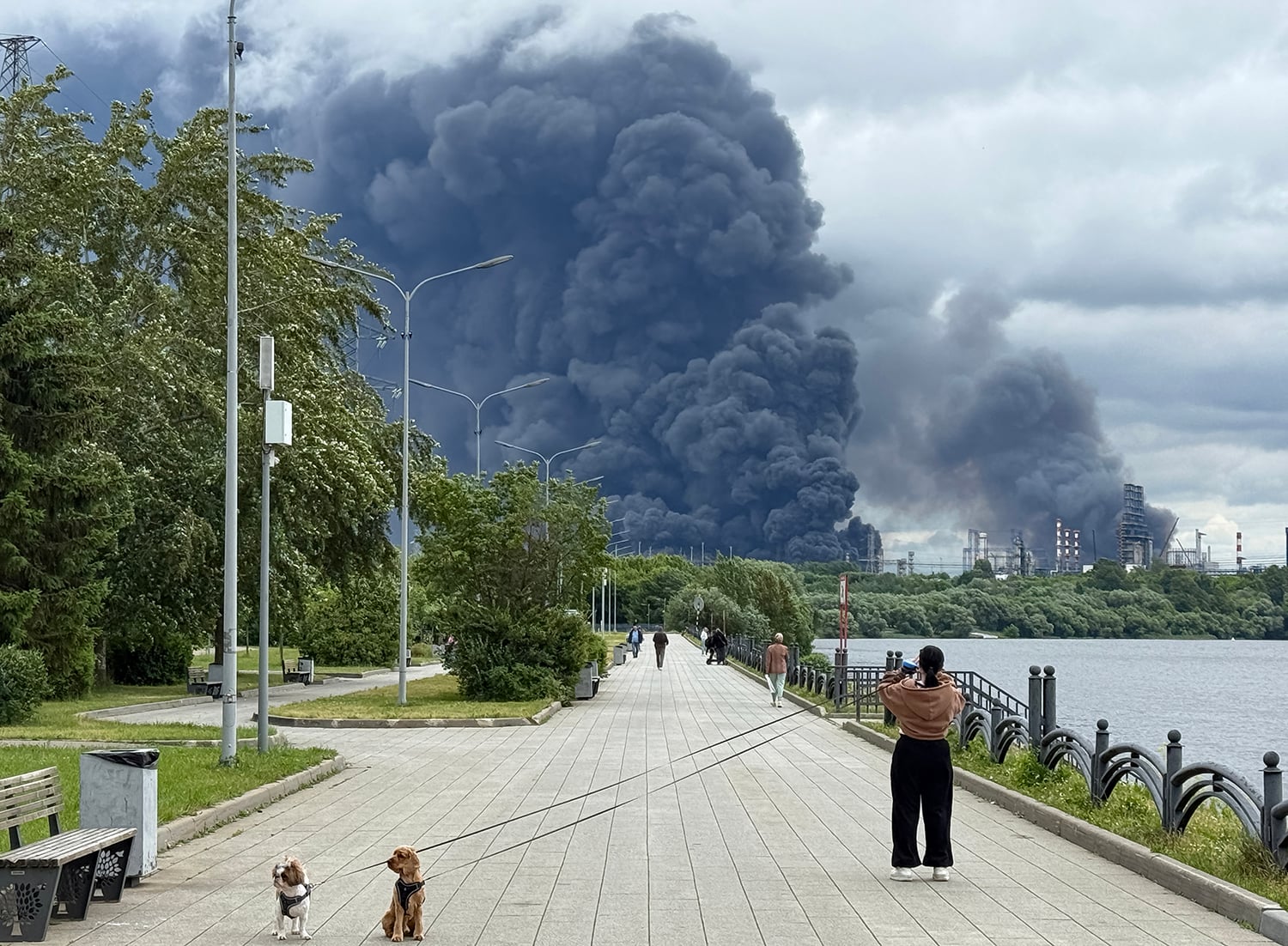 A paved waterfront walkway under an overcast sky. In the foreground, a person walks two small dogs on long leashes, while another person nearby stands looking across the water. In the background across the water, an industrial facility emits an enormous, billowing plume of dark black smoke that stretches high into the cloudy sky.